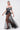 A woman walks confidently in a Parisian-inspired look, wearing a black-and-white off-the-shoulder striped top with a sculptural corset detail and the MARA SHEER POLKA DOT MAXI SKIRT, styled with black heels and gold jewelry against a plain background.