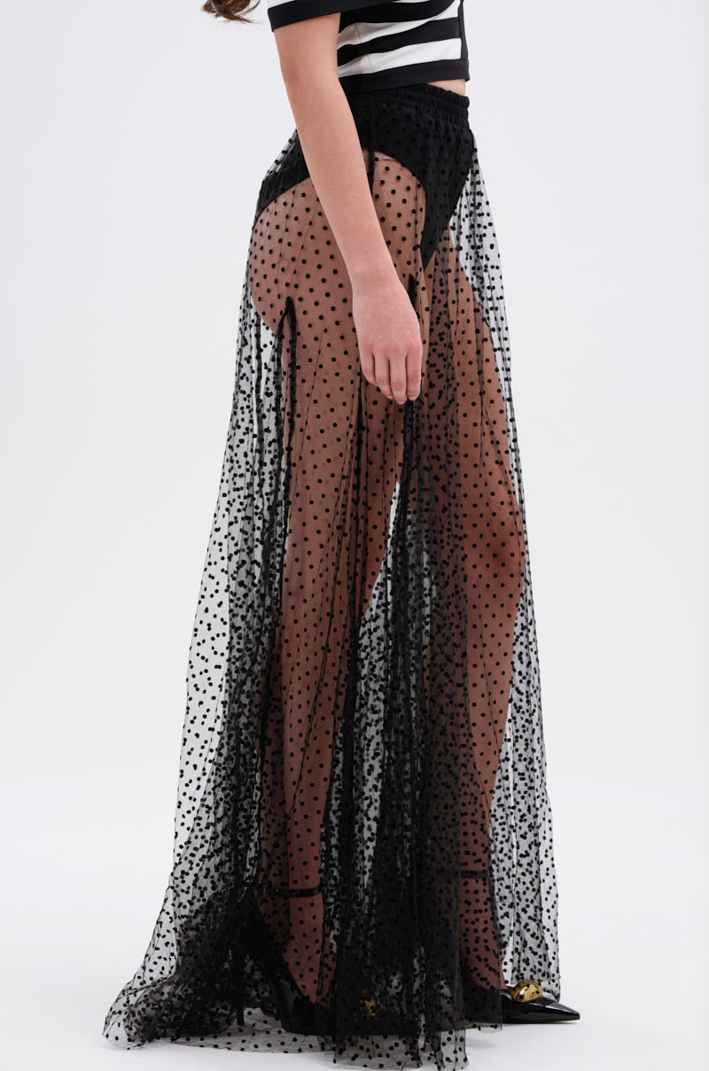 A person models Parisian style in the MARA SHEER POLKA DOT MAXI SKIRT over high-waisted black underwear with a black and white striped crop top and black heels, standing against a plain white background.