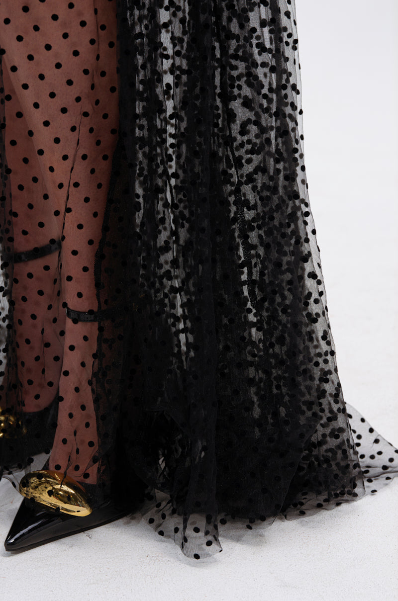 A close-up of the MARA SHEER POLKA DOT MAXI SKIRT draped over legs, paired with a black and gold pointed shoe on a light floor, capturing effortless Parisian style.