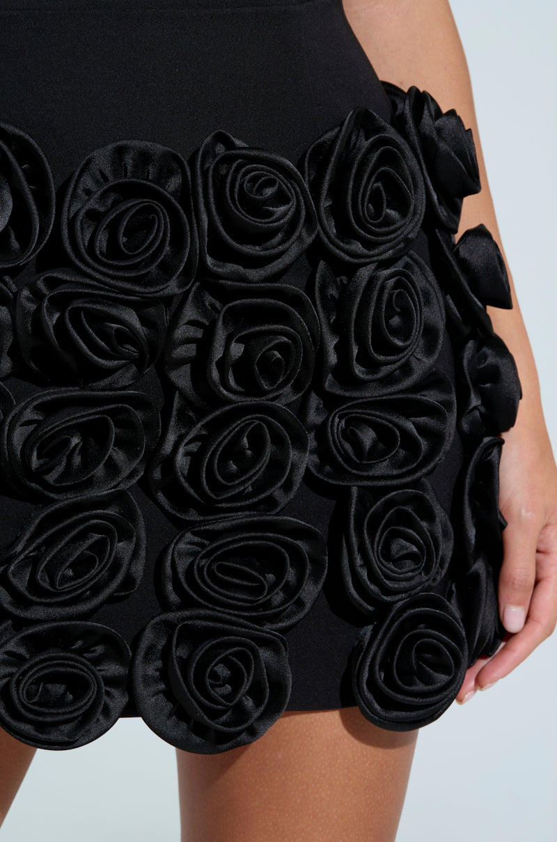 Close-up of a person in the Mind Your Business Mini Dress With Tie, featuring fabric roses that elegantly texture the strapless bodycon silhouette. The simple, light background enhances the floral embellishments on the skirt.