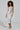 The woman with curly hair stands against a plain background, wearing the dazzling SAMANTHA SEQUIN MIDI DRESS, complete with adjustable tank straps for a perfect bodycon fit. She pairs it elegantly with metallic heels and holds a small silver clutch.