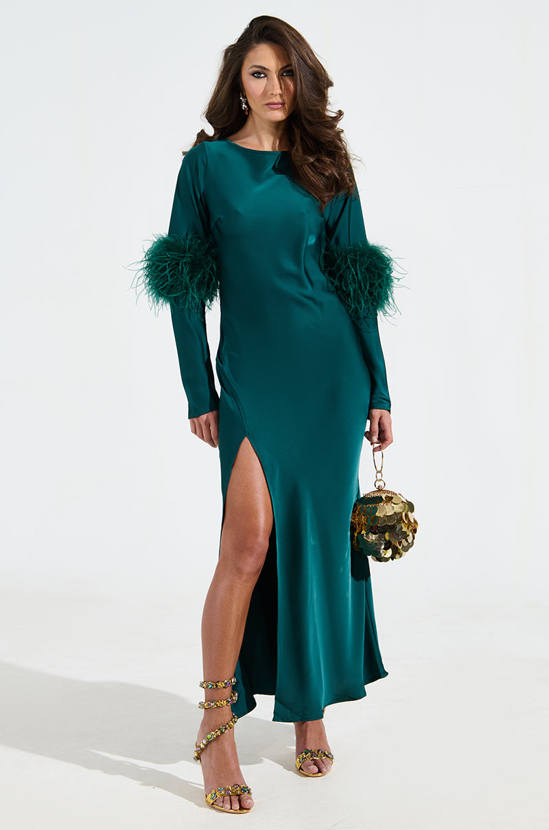 A woman confidently poses in her teal satin Carrie Feather Sleeve Maxi Dress, featuring feathered sleeves and a thigh-high slit. She pairs it with a metallic gold handbag and strappy high-heeled sandals against a light background, showcasing the mermaid-inspired silhouette.