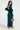 Wearing the CARRIE FEATHER SLEEVE MAXI DRESS, a woman highlights its mermaid-inspired silhouette and satin fabric. The emerald gown has open back feathered sleeves, complemented by a gold sequined handbag and strappy heels against a plain backdrop.