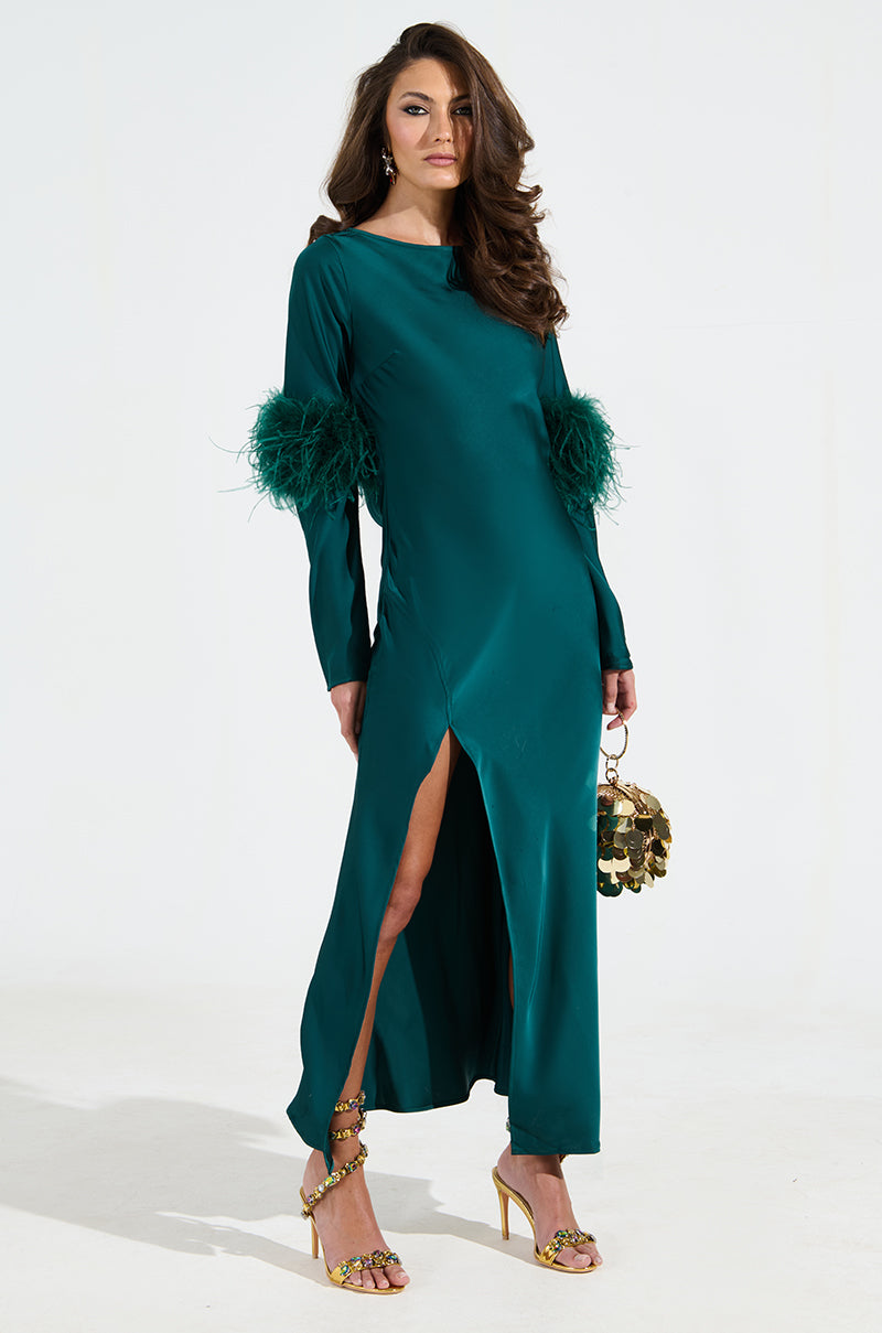 A woman confidently poses in a teal satin CARRIE FEATHER SLEEVE MAXI DRESS, boasting feathered cuffs and a mermaid silhouette with a front slit. She accessorizes with a decorative handbag and gold high heels, her hair styled in loose waves against a neutral background.