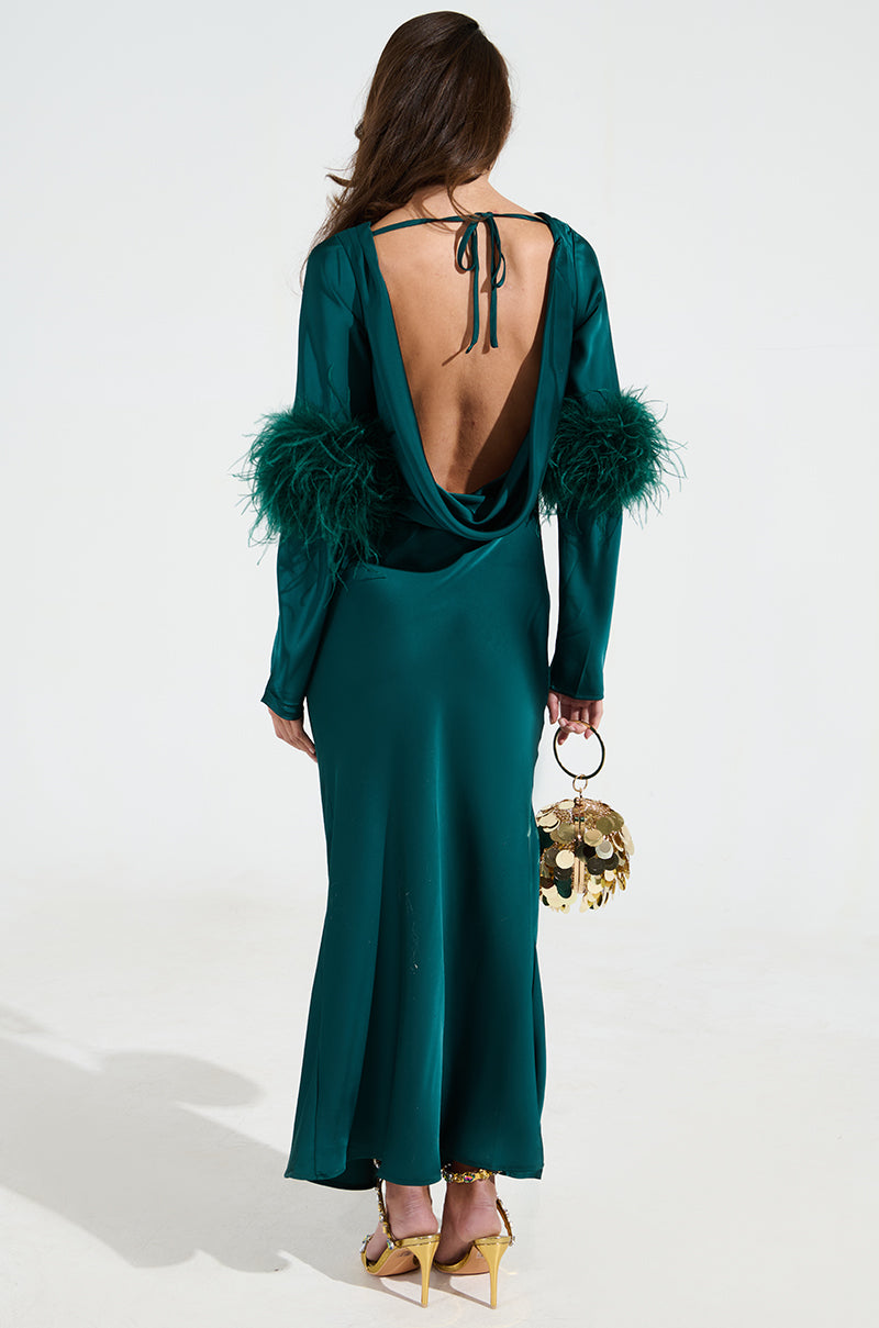 A woman stands gracefully in a satin long, green CARRIE FEATHER SLEEVE MAXI DRESS with a mermaid silhouette and feathered sleeves, carrying a small, round gold handbag. She pairs it with yellow high heels while her hair cascades against a plain background.