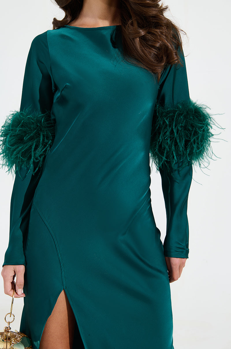 A woman in a sleek teal Carrie Feather Sleeve Maxi Dress, featuring a mermaid silhouette, feathered cuffs, and side slit, holds an ornate handbag. Her long wavy hair cascades over her shoulders, enhancing the dress's elegant satin design.