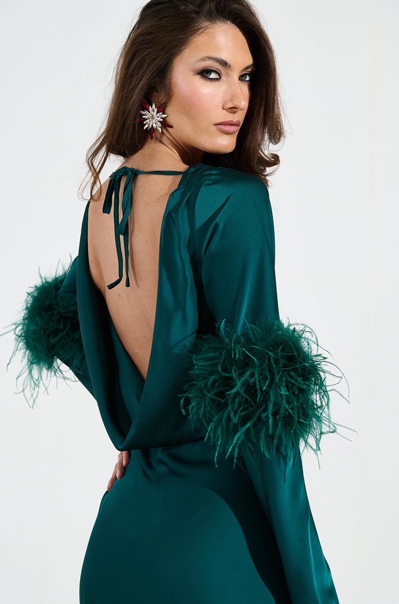 A woman elegantly turns in the CARRIE FEATHER SLEEVE MAXI DRESS, showcasing its mermaid-inspired silhouette. The deep green satin ensemble's open back and feathered sleeves enhance her elegance as statement earrings glint beneath her flowing hair against a plain, light background.