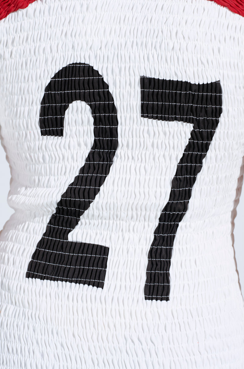 The image showcases a TEAM PLAYER SMOCKED JERSEY DRESS with textured white fabric and a large black "27," evoking a sporty patch. A red section at the top edge complements the color block design.