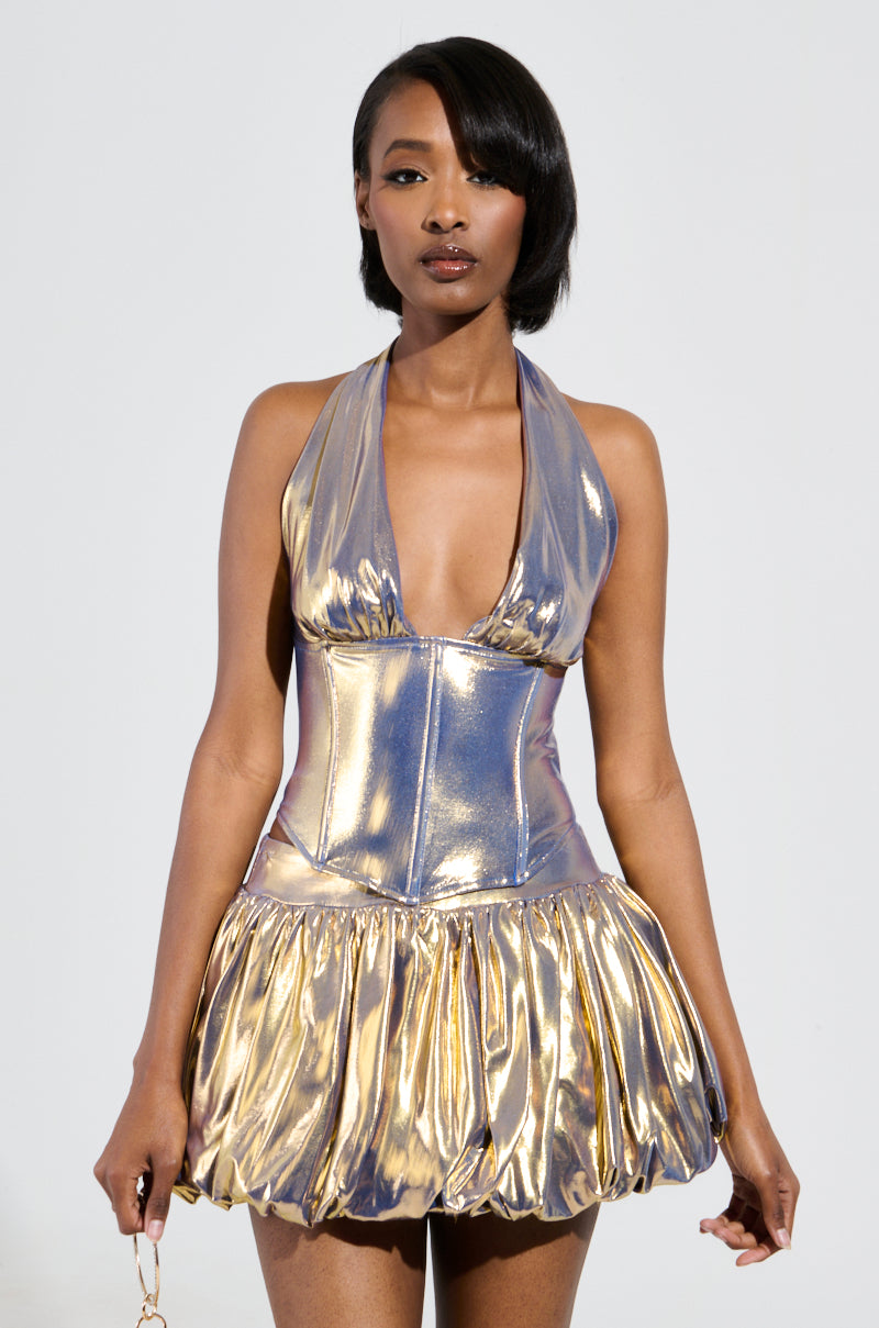 A woman showcasing a glistening, metallic halter dress with a plunging neckline and full skirt stands against a simple backdrop. The style, recalling the BETTER THAN GOLD CORSET TOP by AKIRA Label, includes structural boning and a shimmering knit foundation. Her shoulder-length hair perfectly complements the outfit.