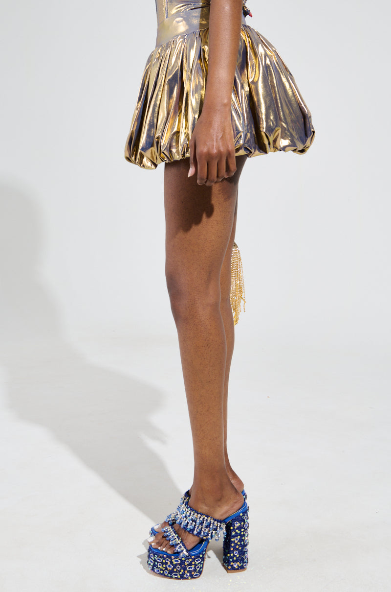 A person is featured wearing the BETTER THAN GOLD BUBBLE MINI SKIRT paired with striking blue high-heeled sandals adorned with colorful embellishments, set against a light background. The image focuses on the lower half, showcasing the ruched construction of the skirt and eye-catching footwear.