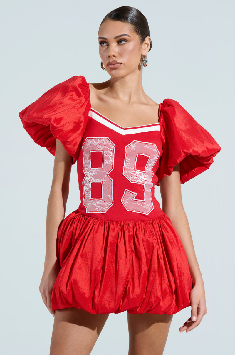 Wearing the "Sorry I'm Late" jersey mini dress in vibrant red with large puffed sleeves and number 89 on the front, one achieves a sporty chic look. The fitted cotton-blend bodice transitions into a voluminous skirt, paired with hoop earrings and an updo against a plain backdrop.