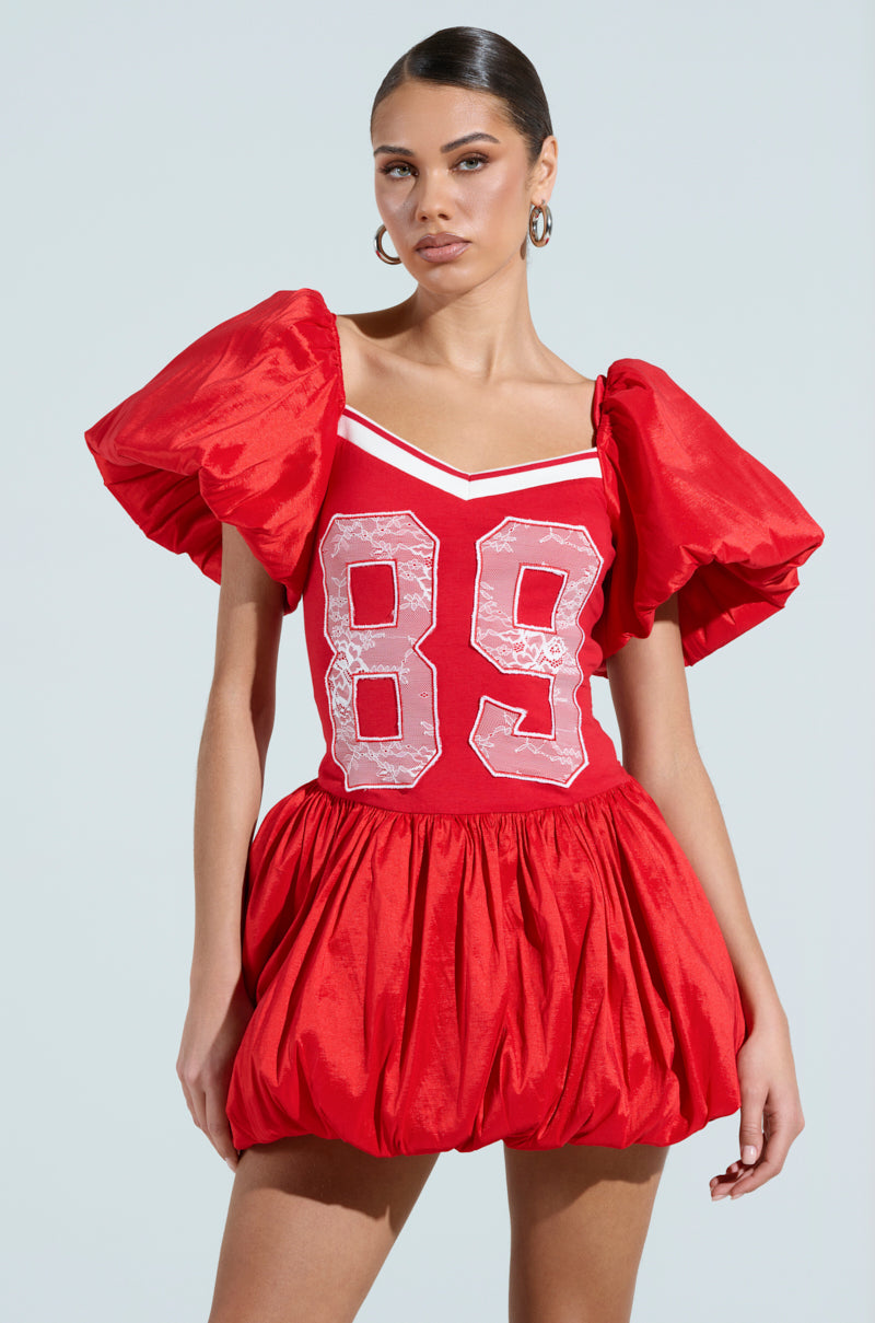 A woman sports the SORRY I'M LATE JERSEY MINI DRESS in red, featuring puffy short sleeves, a skirt, number 89 on the front, and an athletic stripe accent against a plain light blue background.