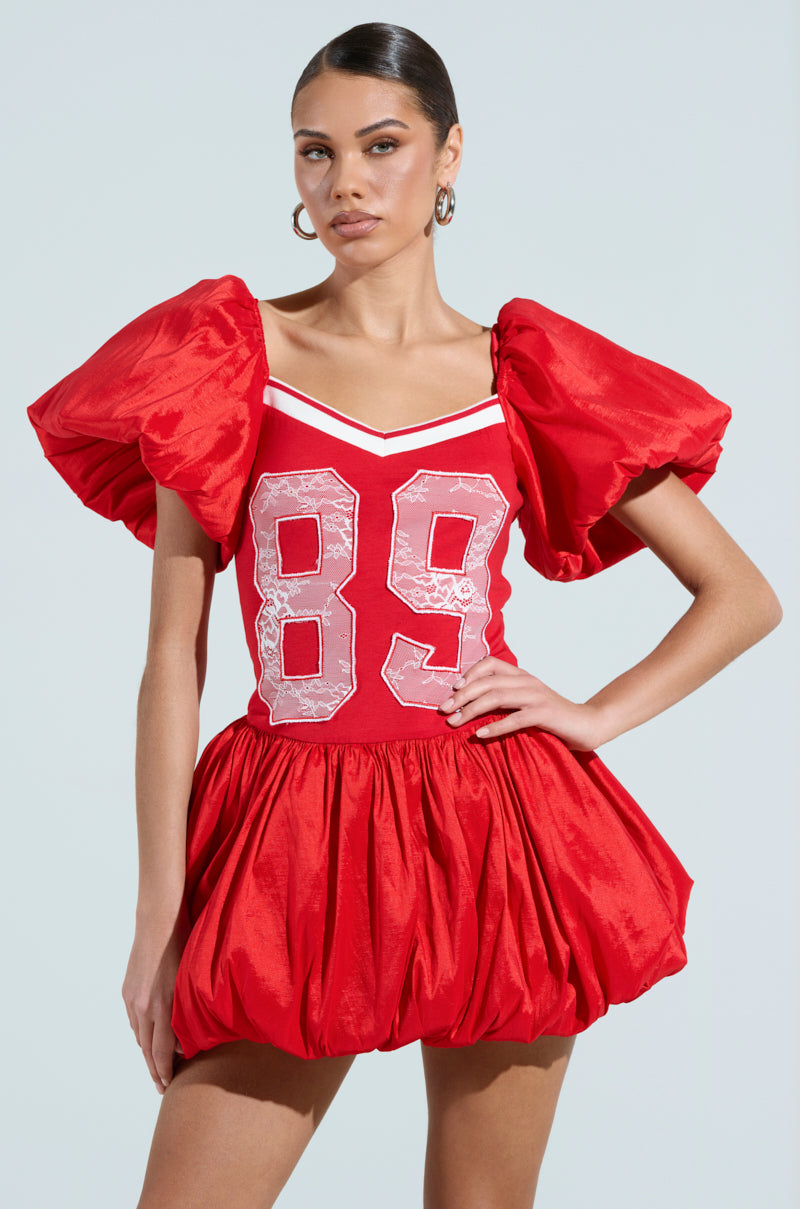 In the SORRY I'M LATE JERSEY MINI DRESS, a sporty chic ensemble, a woman stands confidently. This red masterpiece with puffy sleeves and a bubble skirt showcases the number "89" in bold white on its cotton-blend bodice as she poses against a soft, neutral background.