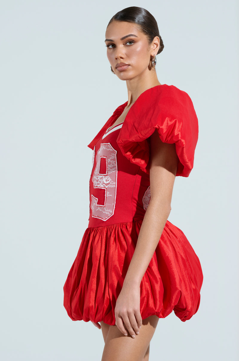 A person models the "SORRY I'M LATE" Jersey Mini Dress in vibrant red, featuring puffed sleeves, a number 9 on the front, and a unique voluminous skirt. They look to the side with sleek hair against a soft light blue background, embodying sporty chic flair.