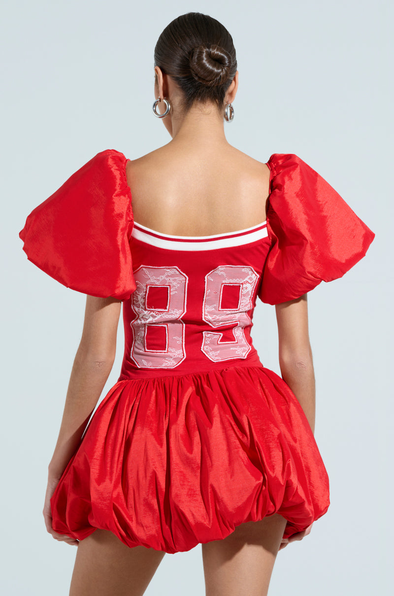 Facing away, a person wears the vibrant SORRY I'M LATE JERSEY MINI DRESS featuring voluminous puff sleeves, a bubble skirt, and the number 89 in bold white on the upper back. Their hair is styled in a neat bun.
