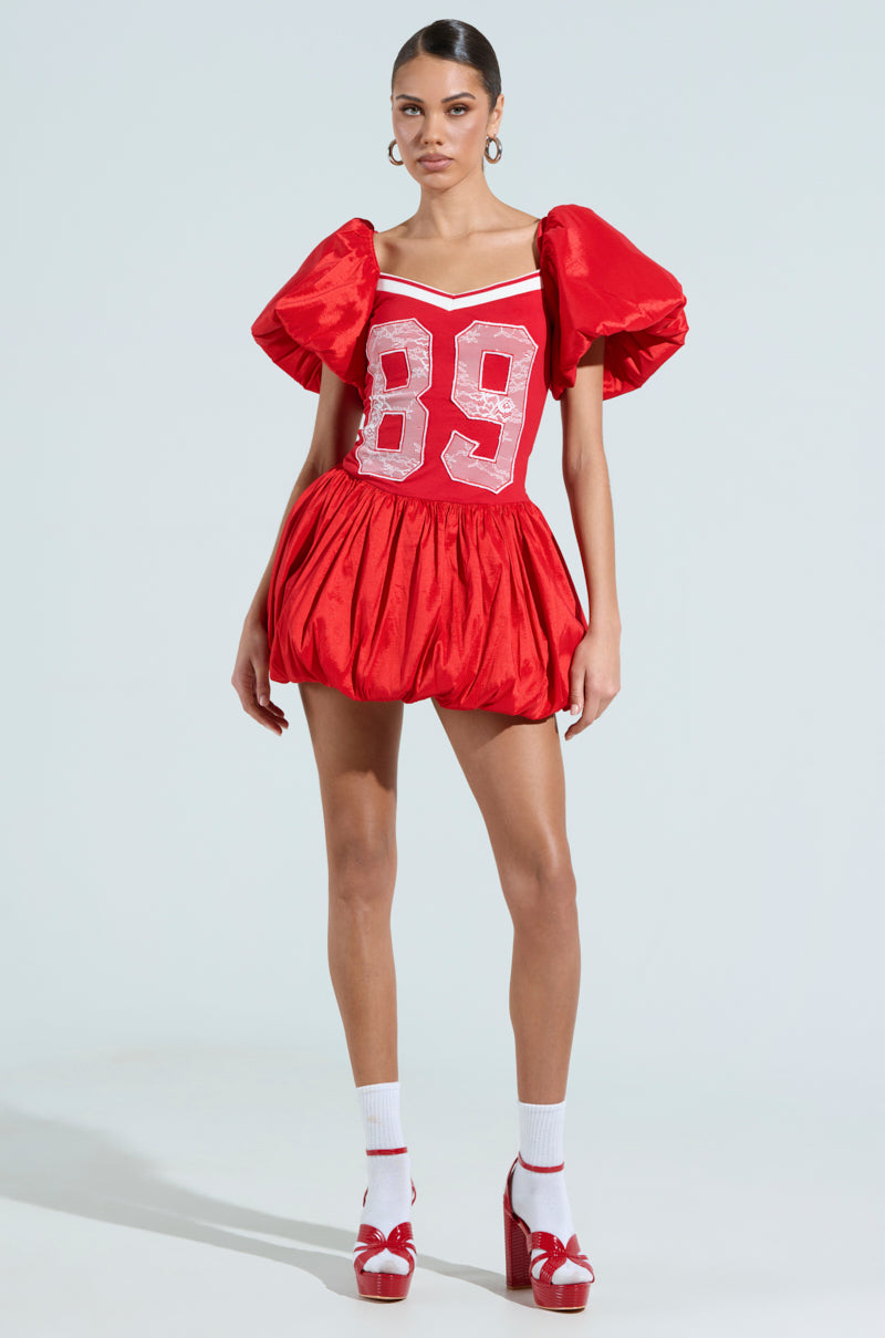 A woman showcases sporty chic in the SORRY I'M LATE JERSEY MINI DRESS, a red cotton-blend piece with puffy sleeves and bold white "89" on the front. She completes the look with white socks, red platform heels, large hoop earrings, and sleekly pulled-back hair.