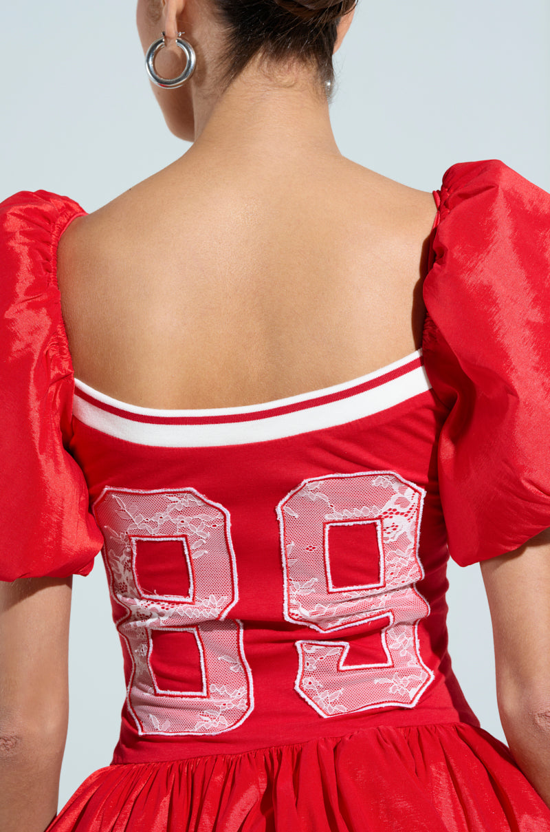 Wearing the SORRY I'M LATE JERSEY MINI DRESS, a sporty chic garment with puffed sleeves and a white stripe near the neckline, they flaunt vibrant red cotton-blend fabric adorned with lace number 89 on the back. Their look is completed with short hair and large hoop earrings.