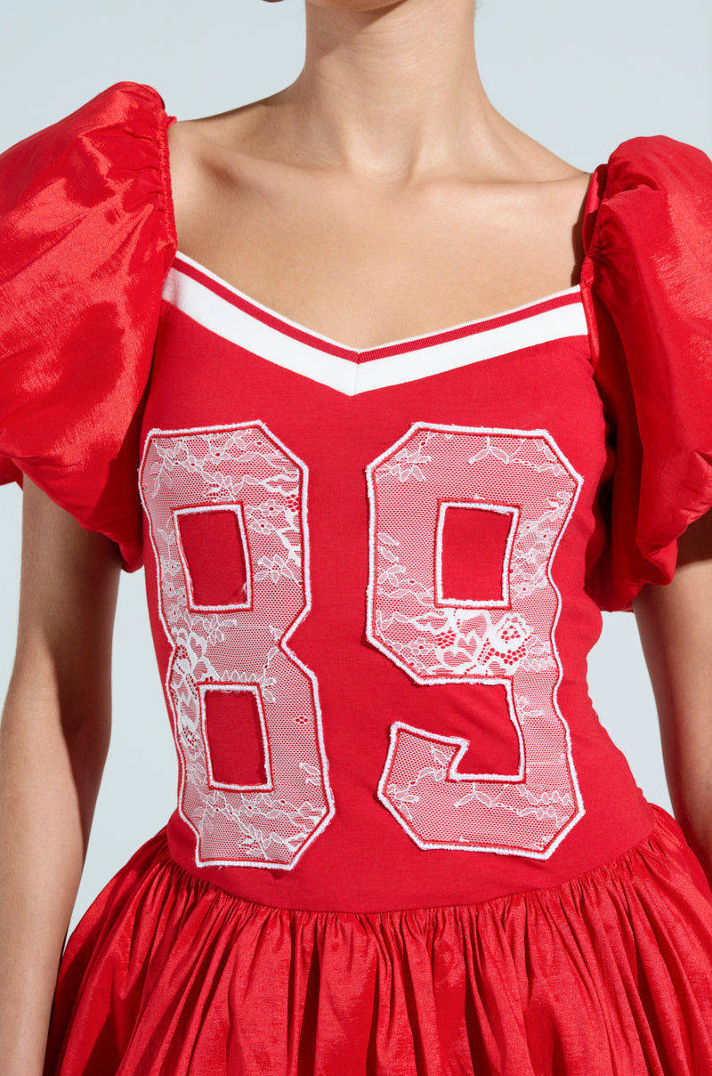 A person is wearing the SORRY I'M LATE JERSEY MINI DRESS in red with puffed sleeves and a flared skirt. The cotton-blend bodice features a large white lace number "89" on the front, set against a plain light blue background.