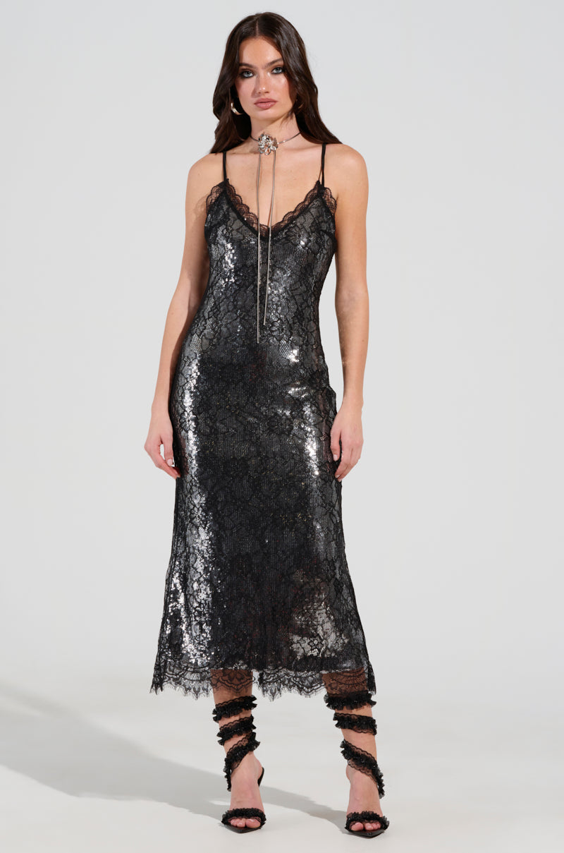 A woman stands against a plain background wearing the TWO IS BETTER THAN ONE lace layered midi dress, which features a black color and shimmering finish. The sleeveless dress also has a scalloped hemline. She complements her look with black strappy heels and a long necklace with a pendant, while her hair is styled in loose waves.