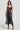 A woman stands against a plain background wearing the TWO IS BETTER THAN ONE lace layered midi dress, which features a black color and shimmering finish. The sleeveless dress also has a scalloped hemline. She complements her look with black strappy heels and a long necklace with a pendant, while her hair is styled in loose waves.