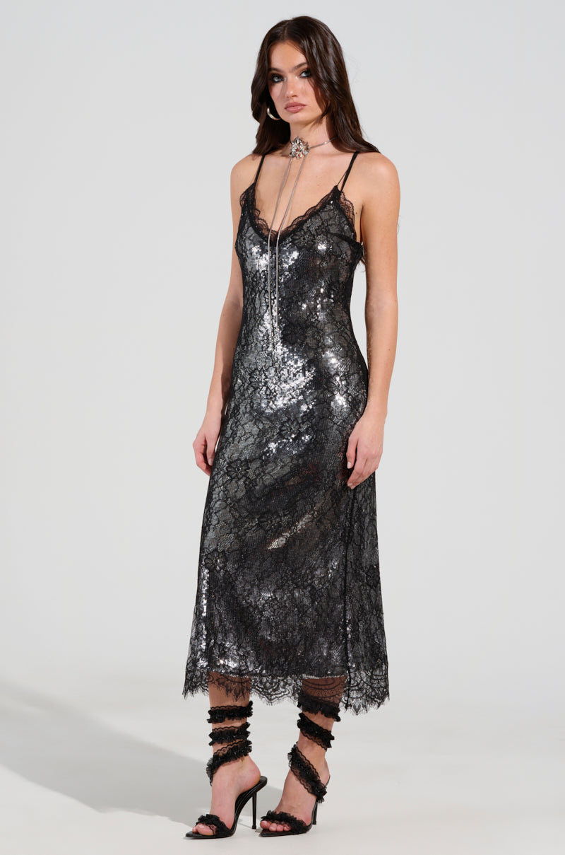 A woman is modeling the TWO IS BETTER THAN ONE LACE LAYERED MIDI DRESS, a sleeveless, knee-length dress in dark silver with lace details. She pairs it with strappy black high heels and a choker complemented by a long necklace, set against a plain light-colored background.
