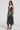 A woman is modeling the TWO IS BETTER THAN ONE LACE LAYERED MIDI DRESS, a sleeveless, knee-length dress in dark silver with lace details. She pairs it with strappy black high heels and a choker complemented by a long necklace, set against a plain light-colored background.