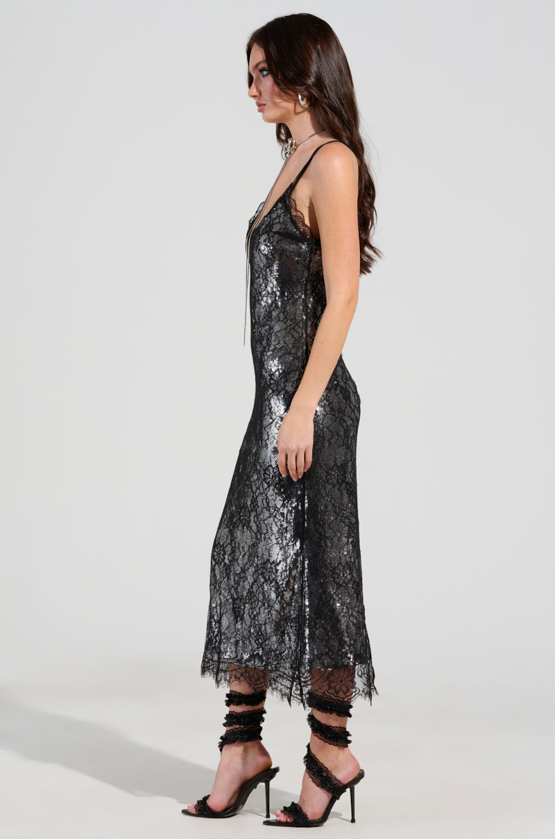 A woman in the TWO IS BETTER THAN ONE lace layered midi dress, which is sleeveless and black, stands in profile against a plain background. She wears high heels, and her long hair cascades down her back.