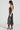 A woman in the TWO IS BETTER THAN ONE lace layered midi dress, which is sleeveless and black, stands in profile against a plain background. She wears high heels, and her long hair cascades down her back.