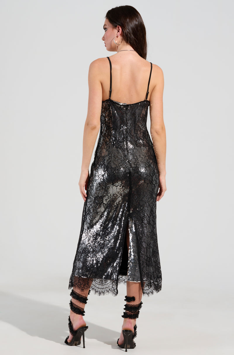 A woman in the elegant TWO IS BETTER THAN ONE LACE LAYERED MIDI DRESS, featuring a shiny, sheer mesh with sequin embellishments, stands with her back to the camera. The dress is designed with thin straps and a back slit. She completes her look with strappy, embellished high heels.