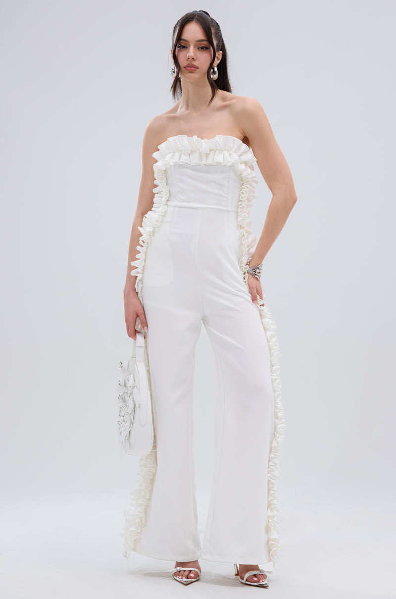 A woman stands against a plain light background, wearing the MONICA RUFFLED JUMPSUIT IN WHITE with a strapless neckline and wide-leg silhouette, paired with a white handbag and white heeled sandals.