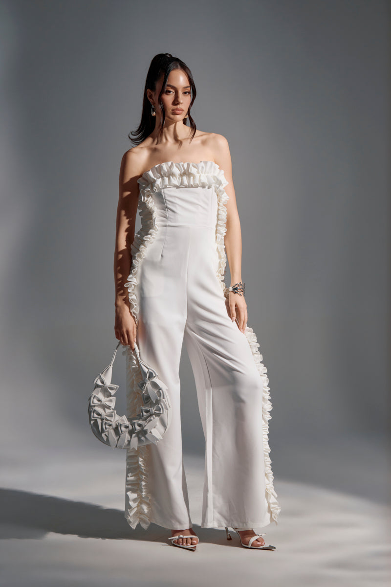 A woman stands in a studio wearing the MONICA RUFFLED JUMPSUIT IN WHITE, featuring a strapless neckline and wide-leg silhouette. She holds a white ruffled handbag, wears heeled sandals, and has her hair in a high ponytail.