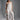 A woman stands in a studio wearing the MONICA RUFFLED JUMPSUIT IN WHITE, featuring a strapless neckline and wide-leg silhouette. She holds a white ruffled handbag, wears heeled sandals, and has her hair in a high ponytail.