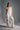 A woman stands in a studio wearing the MONICA RUFFLED JUMPSUIT IN WHITE, featuring a strapless neckline and wide-leg silhouette. She holds a white ruffled handbag, wears heeled sandals, and has her hair in a high ponytail.