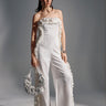 A woman stands in a studio wearing the MONICA RUFFLED JUMPSUIT IN WHITE, featuring a strapless neckline and wide-leg silhouette. She holds a white ruffled handbag, wears heeled sandals, and has her hair in a high ponytail.
