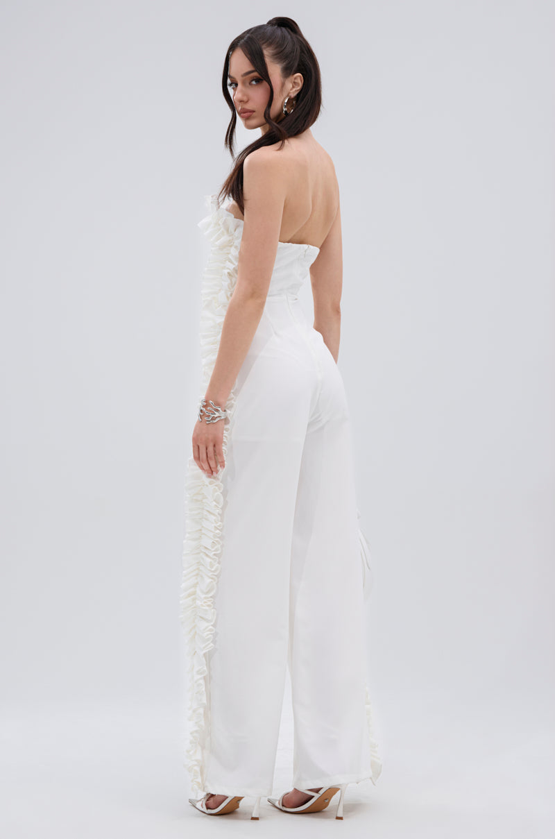 A woman stands sideways, looking over her shoulder in the MONICA RUFFLED JUMPSUIT IN WHITE, featuring a strapless neckline and wide-leg silhouette. She pairs it with white high heels, loose waves, and chic bracelets.