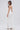 A woman stands sideways, looking over her shoulder in the MONICA RUFFLED JUMPSUIT IN WHITE, featuring a strapless neckline and wide-leg silhouette. She pairs it with white high heels, loose waves, and chic bracelets.