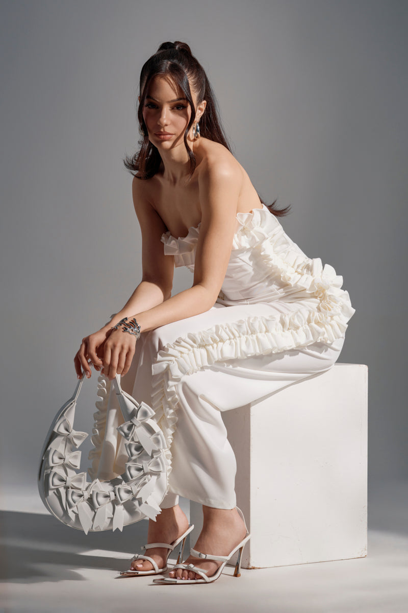 A woman in the MONICA RUFFLED JUMPSUIT IN WHITE, featuring a strapless neckline and wide-leg silhouette, sits on a white cube holding a ruffled white handbag. She completes the look with white high-heeled sandals and loose wavy hair.