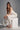 A woman in the MONICA RUFFLED JUMPSUIT IN WHITE, featuring a strapless neckline and wide-leg silhouette, sits on a white cube holding a ruffled white handbag. She completes the look with white high-heeled sandals and loose wavy hair.