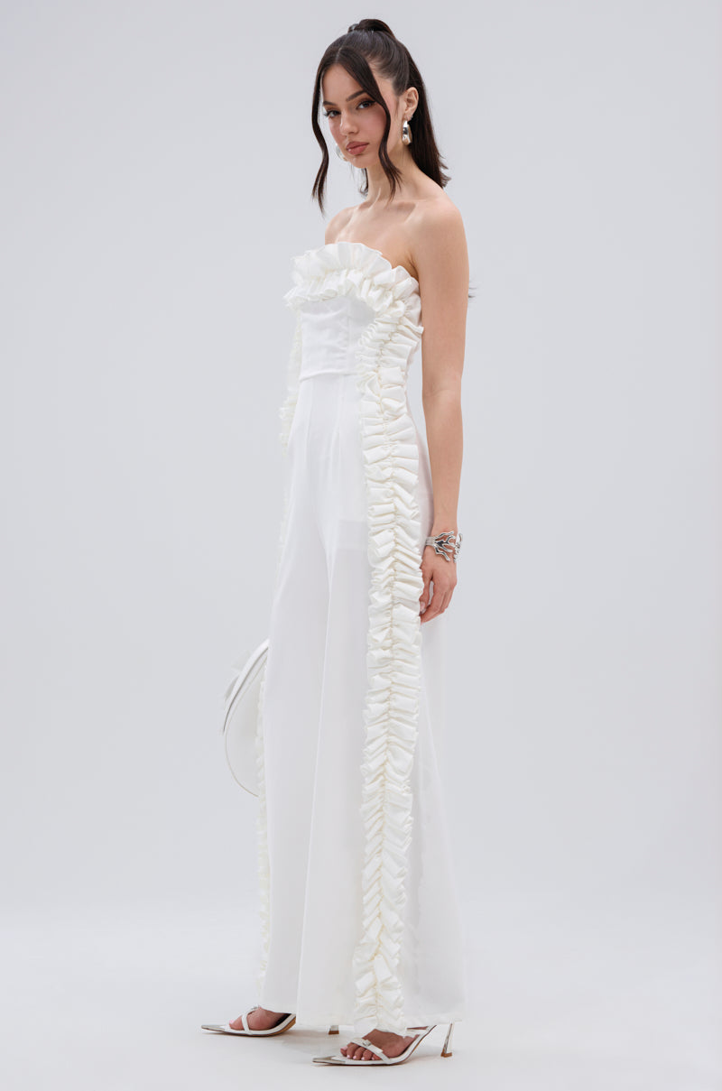 A woman stands against a plain background wearing the MONICA RUFFLED JUMPSUIT IN WHITE, featuring a strapless neckline and ruffled trim down the sides. She pairs it with a white clutch, white heels, silver jewelry, and loose waves in her long dark hair.
