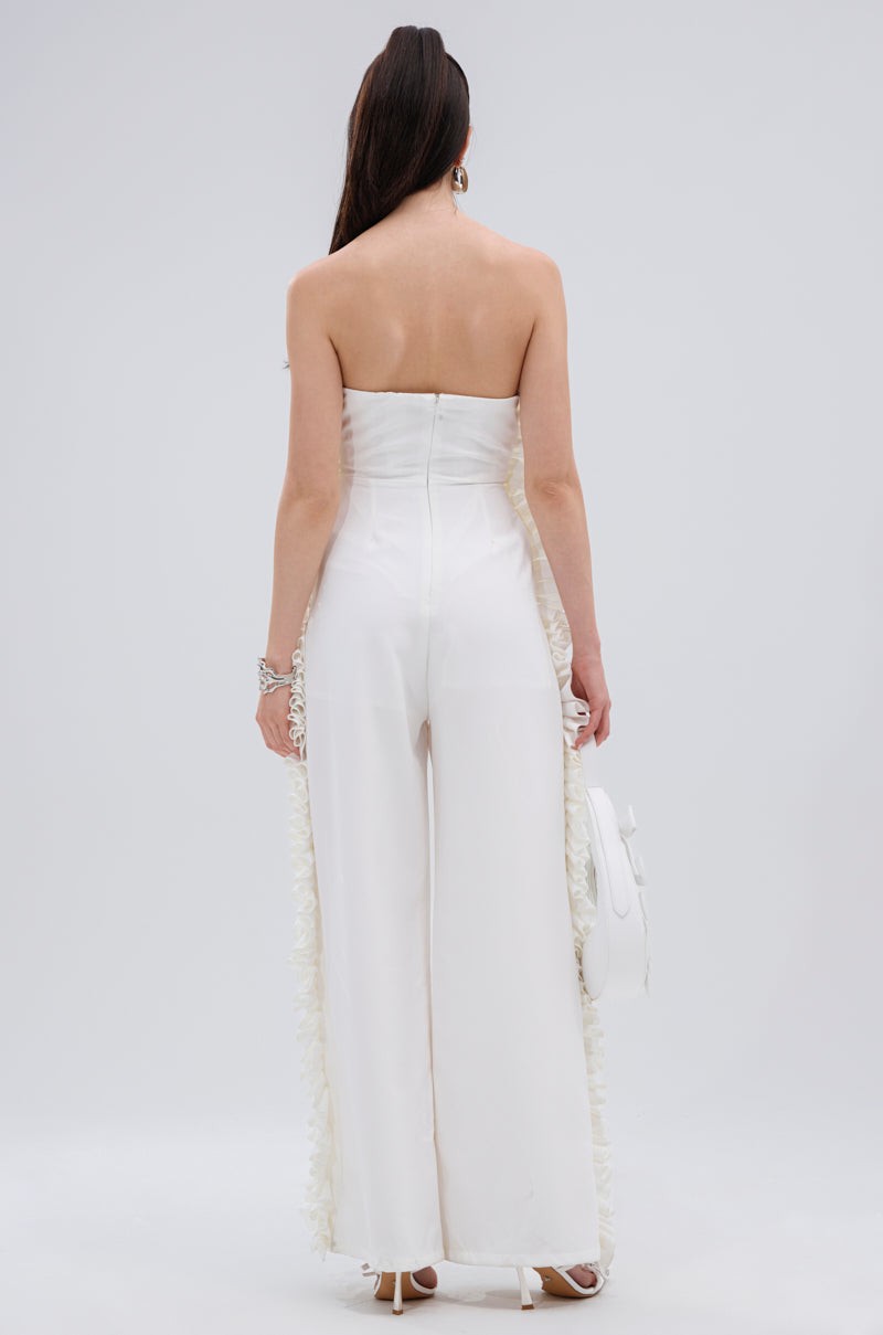 A woman stands facing away, wearing the MONICA RUFFLED JUMPSUIT IN WHITE, featuring a strapless neckline and wide-leg silhouette. She carries a white handbag, with her long dark hair styled straight down her back.