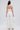 A woman stands facing away, wearing the MONICA RUFFLED JUMPSUIT IN WHITE, featuring a strapless neckline and wide-leg silhouette. She carries a white handbag, with her long dark hair styled straight down her back.