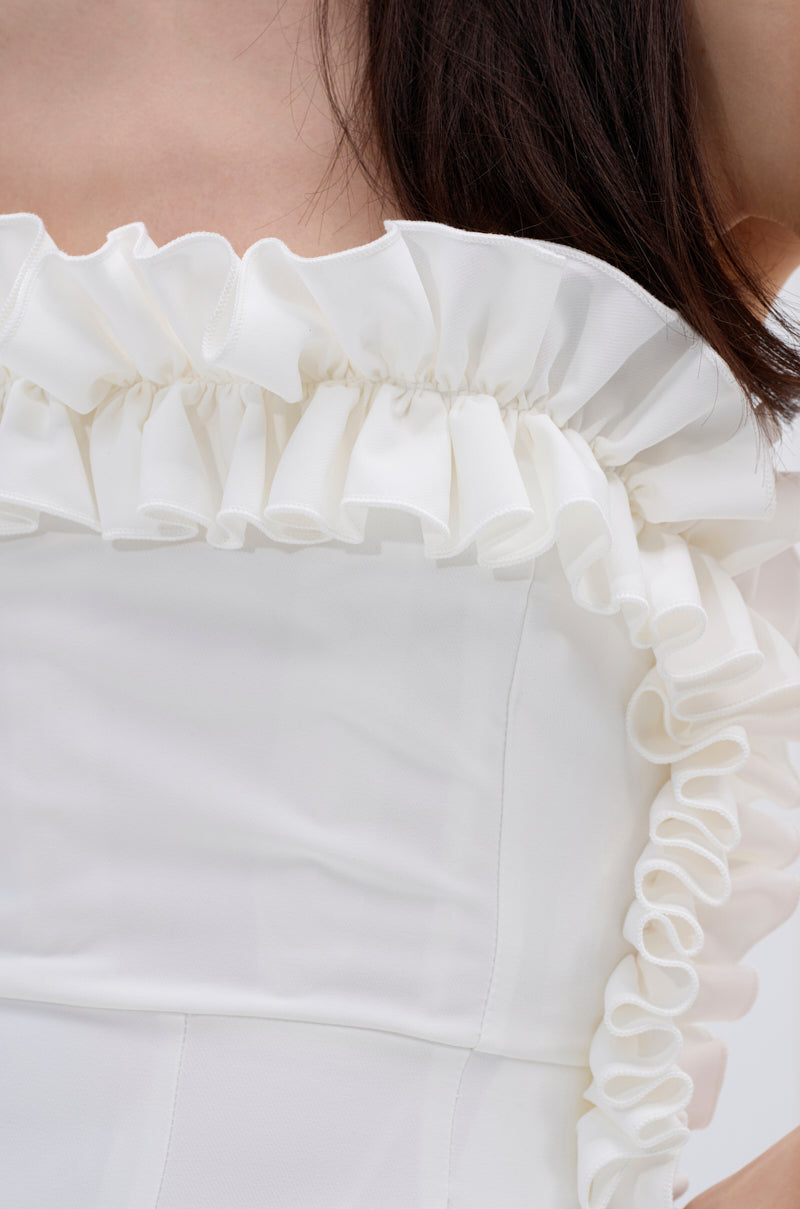 Close-up of a person wearing the MONICA RUFFLED JUMPSUIT IN WHITE, featuring an off-the-shoulder design with delicate ruffle detailing along the neckline and sleeves. The soft, slightly sheer fabric complements their dark hair draped over one shoulder.