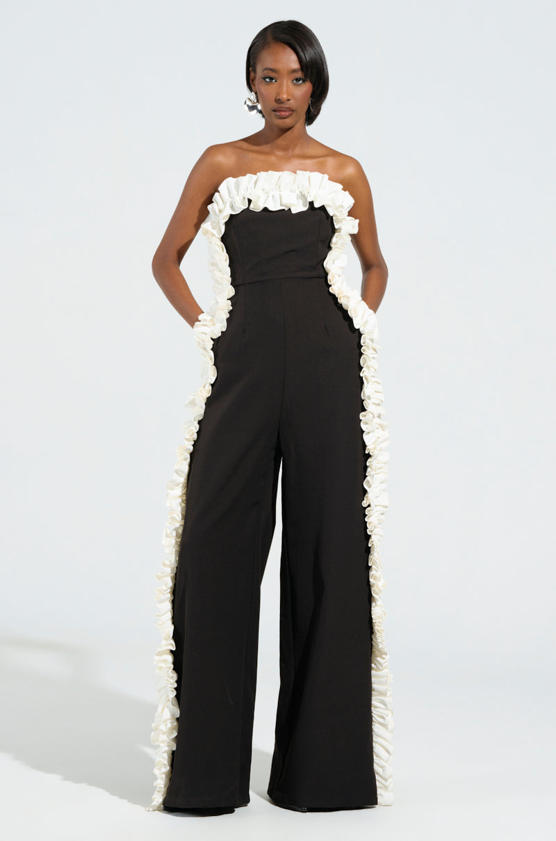 A woman stands confidently in the MONICA RUFFLED JUMPSUIT IN BLACK, elegantly highlighted by white ruffles along the top and sides. The strapless design adds a touch of sophistication against the plain light background.