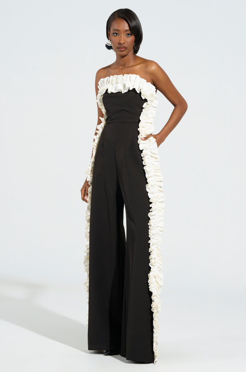 A model exudes confidence in the MONICA RUFFLED JUMPSUIT IN BLACK, featuring a striking strapless design highlighted by white ruffles adorning the neckline, sides, and hem. The wide-leg jumpsuit drapes gracefully as she poses against a plain white background.