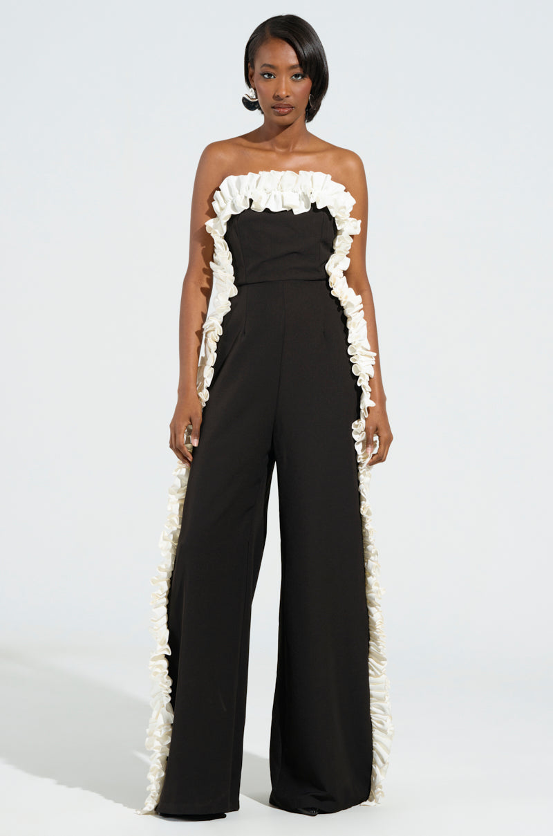 A woman stands confidently in the MONICA RUFFLED JUMPSUIT IN BLACK, showcasing its strapless design with wide legs and elegant white trim along the neckline and sides. Her hair is styled sleekly, and she wears a neutral expression against a light background.