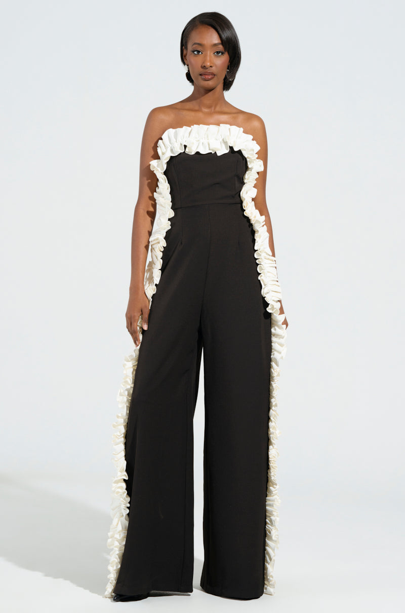 A woman exudes elegance in the MONICA RUFFLED JUMPSUIT IN BLACK, showcasing a strapless design with white ruffled trim adorning the top and sides. Her shoulder-length dark hair enhances her confident gaze against a light background, beautifully highlighting the chic silhouette of this wide-leg jumpsuit.