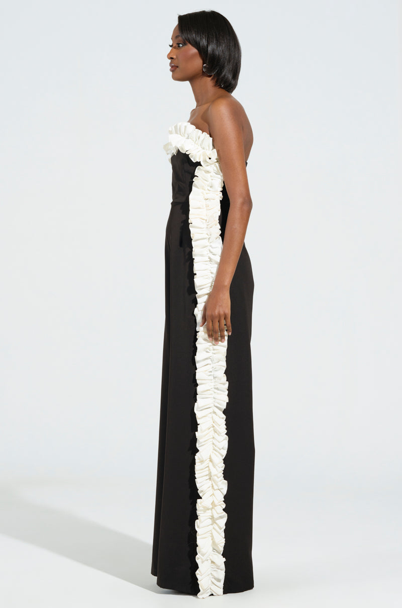 A woman stands in profile wearing the MONICA RUFFLED JUMPSUIT IN BLACK, featuring a prominent white ruffle detail running vertically. Her hair is styled straight, set against a light neutral background.