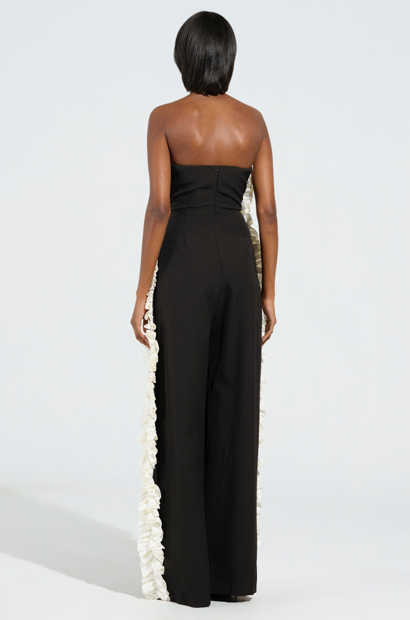 A person stands with their back to the camera, showcasing the Monica Ruffled Jumpsuit in black. This strapless wide-leg jumpsuit features a stylish white ruffle detail along the side and contrasts beautifully against a plain, light-colored background.