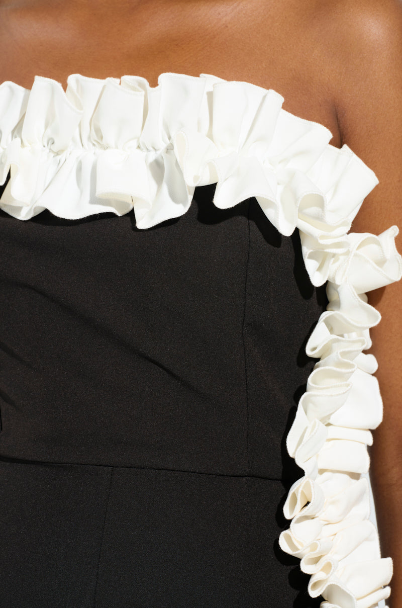 A close-up shot features an individual wearing the MONICA RUFFLED JUMPSUIT IN BLACK, showcasing a strapless design with charming black ruffles and a white ruffled trim along the top edge. The textured ruffles beautifully contrast with the smooth fabric, emphasizing its elegant design while revealing some skin above the jumpsuit.