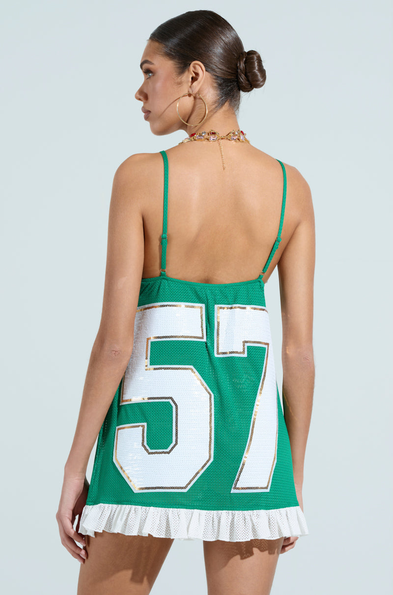 Wearing hoop earrings and a necklace, the person is dressed in the Touchdown Sequin Jersey Dress in green. It features a white number 57 on the back, thin straps, a white ruffled hem, and a V-neckline against a light blue background.